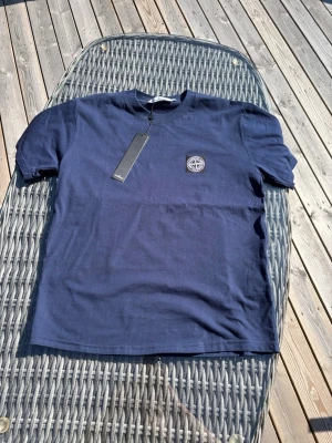 Mörkblå Stone Island t-shirt - Snygg mörkblå t-shirt från Stone Island med klassisk logopatch på bröstet. T-shirten har rund hals och korta ärmar, tillverkad i mjuk bomull för en skön känsla. Perfekt för dig som gillar streetwear och vill ha en stilren look.