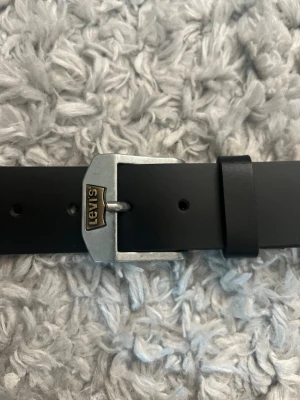 Svart skärp från Levi's i skinn - Stilrent svart skärp från Levi's i äkta skinn med klassiskt silverfärgat spänne och logga på spännet. Perfekt accessoar för att lyfta din outfit med en tidlös touch.