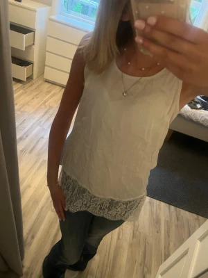 Vit spetslinne från Define - Snyggt vitt långt linne från Define med spetsdetalj längst ner. Luftigt material och ärmlös design, perfekt till jeans eller kjol. Enkel och stilren look som passar till många outfits.