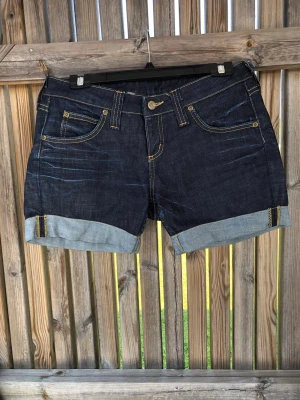 Mörkblå jeansshorts från Lee - Säljer ett par mörkblå jeansshorts från Lee med klassisk femficksdesign och uppvikta benslut. Shortsen har kontrastsömmar och Lee-logga bak på linningen. Perfekta för varma dagar och ger en avslappnad look.