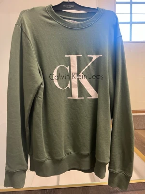 Grön sweatshirt från Calvin Klein - Stilren grön sweatshirt från Calvin Klein Jeans med stort vitt CK-tryck och svart text på bröstet. Tröjan har rund halsringning, ribbade muddar och är tillverkad i mjuk bomullsblandning. Perfekt för en avslappnad och trendig look.