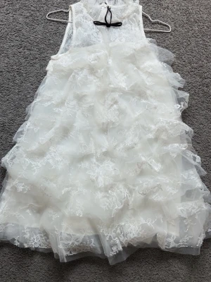 Vit volangklänning med spetsdetaljer - Superfin vit klänning med flera lager volanger och spetsdetaljer. Ärmlös modell med söt rosett i nacken och luftig känsla. Perfekt för dig som vill sticka ut med en romantisk look.