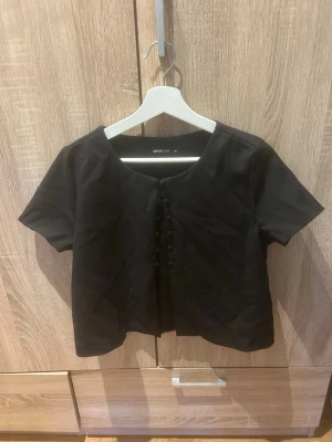 Svart kortärmad topp - Stilren svart topp från Gina Tricot med korta ärmar och snörningsdetalj framtill. Toppen har en något croppad passform och rundad halsringning. Perfekt att matcha med både jeans och kjol.