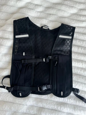 Svart löparväst med reflexdetaljer - Säljer en svart löparväst med flera praktiska fickor framtill och baktill. Västen har justerbara remmar och reflexdetaljer för ökad synlighet. Perfekt för löpning eller träning utomhus när du vill ha med dig småsaker. Onesize. 