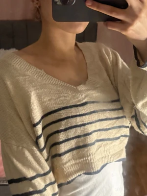 Beige stickad tröja med blå ränder - Säljer en beige stickad tröja med mörkblå horisontella ränder. Tröjan har en djup v-ringning, är croppad och har långa ärmar med ribbade muddar. Perfekt för dig som gillar en avslappnad och trendig look.