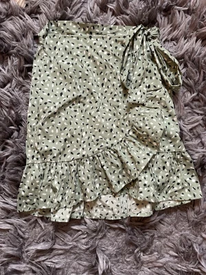 Grön omlottkjol med volang - Superfin grön-mönstrad kort omlottkjol med svartvitt prickigt mönster och volangkant. Kjolen har en knytning i midjan och är gjord i ett lätt och skönt material, perfekt för varmare dagar. Passar dig som vill ha en trendig och bekväm look. Den perfekt till sommaren!!☀️Skriv om ni villnha bilder på💕 Lägg gärna bud!