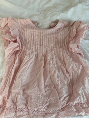 Ljusrosa blus med volangärmar - Söt ljusrosa blus med volangärmar och broderade små blommor. Blusen har rund halsringning, stor i storlek för den passar mig som har xs. veckad framsida och spetsdetalj längst ner. Tillverkad i ett tunt, luftigt tyg som känns romantiskt och somrigt.