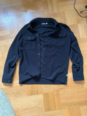 Svart skjorta från Only & Sons - Stilren svart skjorta från Only & Sons med klassisk krage, två bröstfickor med lock och knappar framtill. Skjortan har långa ärmar och en rak passform. Perfekt för en clean och enkel look.