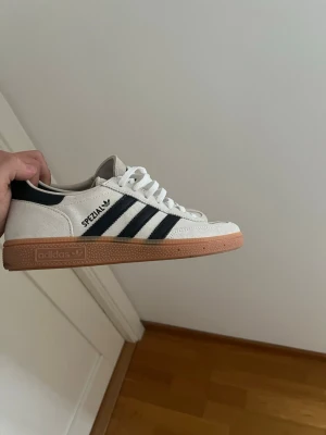 Adidas Spezial vita sneakers - Snygga Adidas Spezial sneakers i vitt mocka med klassiska svarta ränder och brun gummisula. Skon har låg profil och vita skosnören. Perfekt för dig som gillar retrostil och streetwear.