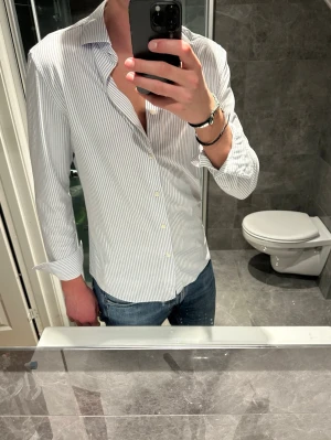 Vit smalrandig skjorta - Klassisk vit skjorta med tunna grå ränder och lång ärm. Skjortan är i riktigt bra skick utan några defekter. Jag på bilden är 188cm och bär storlek M. Hör av er vid frågor:)