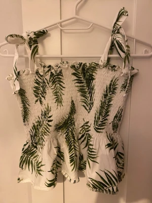 Vit linnetopp med grönt palmtryck - Söt vit linnetopp med grönt palmbladsmönster och smockad överdel. Axelbanden knyts på axlarna för en personlig touch och toppen har volangkant nedtill. Perfekt för varma dagar och somriga outfits.