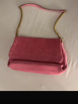 Rosa axelremsväska i mocka - Snygg rosa handväska i mjuk mocka med guldfärgad kedjerem och axelparti i matchande rosa material. Väskan har en mjukt formad design och lock, perfekt för dig som vill ha en trendig accessoar med lyxig känsla.