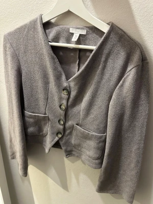 Grå kofta från hm - Säljer en grå kort kofta från H&M med v-ringning och fyra snygga knappar framtill. Koftan har två stora fickor och är gjord i ett mjukt, stickat material. Perfekt att slänga över en topp för en chill look. Tröjan är sparsamt använt och har inga defekter
