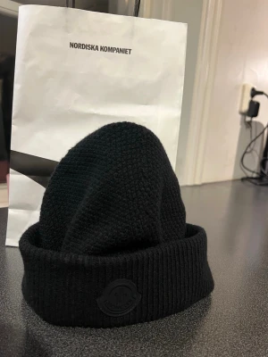 Svart Moncler mössa med logga - Säljer en svart ribbstickad mössa från Moncler med uppvikt kant och diskret svart Moncler-logga framtill. Mössan är i mjukt och värmande material, perfekt för kalla dagar och ger en clean streetwear-look. Köpt på NK i Göteborg, finns kvitto. Nypris 3199 mitt pris 1000