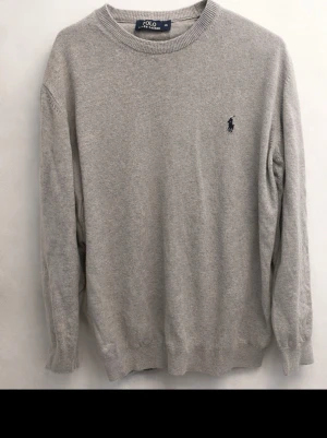 Grå stickad tröja Polo Ralph Lauren - Klassisk grå stickad tröja från Polo Ralph Lauren med rund halsringning och diskret broderad logga på bröstet. Tröjan har ribbade muddar vid ärmslut och nederkant. Perfekt för en stilren och avslappnad look.