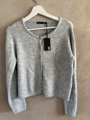Ljusgrå kofta från 157 - Mysig ljusgrå kofta från 157 med rund halsringning och knappar framtill. Koftan är stickad i mjukt material och har långa ärmar samt ribbade muddar vid ärmslut och nederkant. Enkel och stilren design som passar till det mesta.