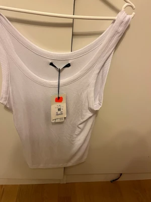 Vit ribbad ärmlös linnetopp - Säljer en fräsch vit linnetopp med bred ribbad kant runt halsen. Toppen är ärmlös och har en clean, enkel design som passar perfekt till jeans eller kjol. Materialet känns mjukt och stretchigt för en bekväm passform.