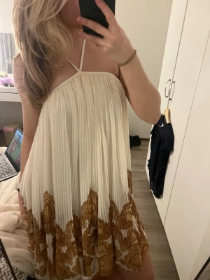 H&M klänning - Säljer en luftig kortklänning i plisserat tyg med tunna axelband och omlottkänsla. Klänningen är vit med beige broderade detaljer nedtill och har en flowy passform som ger en bohemisk vibe. Perfekt för varma dagar och sommarens alla tillfällen. Storlek XS