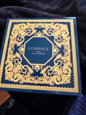 Versace Eros Eau de Toilette presentset - Lyxigt presentset från Versace med Eros Eau de Toilette och tillhörande duschgel. Förpackningen är mörkblå med eleganta gulddetaljer och klassiskt Versace-mönster. Flaskan är mörkblå med Versace-logga och guldtext, och tuben matchar i färg och design.