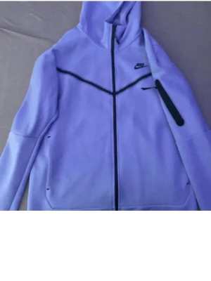 Ljuslila Nike hoodie med dragkedja -   SKICKA MEDDELANDE FÖR ATT BIDDA. Nike hoodie i ljuslila med hel dragkedja framtill och svarta detaljer. Hoodien har en huva, två fickor och en svart linje över bröstet som ger en sportig vibe. Tillverkad i mjukt material som känns skönt mot huden. Perfekt för en avslappnad streetstyle.