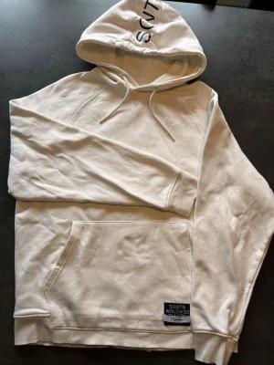 Vit hoodie från SGNTR Worldwide - Snygg vit hoodie från SGNTR Worldwide med svart tryck på huvan och ryggen. Hoodien har känguruficka framtill, dragsko i huvan och ribbade muddar. Perfekt för en avslappnad streetwear-look.