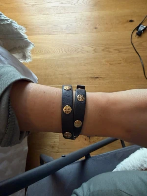 Svart läderarmband från Tory Burch - Snyggt svart läderarmband från Tory Burch med dubbla remmar och guldpläterade runda detaljer med logotyp. Armbandet har ett stilrent spänne i guld och passar perfekt för dig som gillar klassiska accessoarer med en twist. Tillkommer en dustbag. 