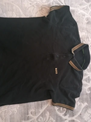 Svart pikétröja från BOSS - Svart pikétröja från BOSS med broderad logga på bröstet. Tröjan har klassisk krage och knäppning framtill samt kontrastfärgade detaljer i beige på krage och ärmslut. Perfekt för en stilren look.