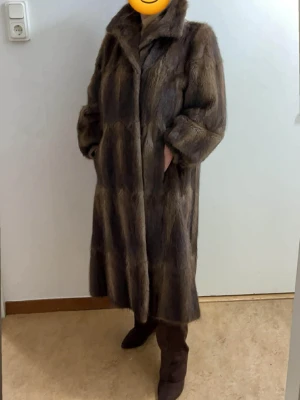 Lång brun minkpäls kappa - Säljer en lång, brun minkpäls kappa med bred krage och dolda fickor. Kappan har en rak passform och stänger framtill. Perfekt för dig som vill ha en lyxig och varm look under kalla dagar. Mössan ingår
