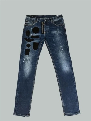 DSQUARED2 Jeans - DSQUARED2 blåa jeans med slitningar och ljus fading på låren. Designen har svarta patch-detaljer på ena låret och en modern, urban stil.