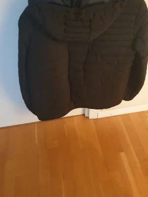 Svart pufferjacka från Tommy Hilfiger - Snygg svart pufferjacka från Tommy Hilfiger med huva och dragkedja framtill. Jackan har diskreta logodetaljer på bröstet och ärmen samt två sidofickor. Perfekt för kalla dagar och ger en stilren look.