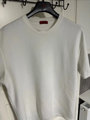 Vit t-shirt från Hugo Boss - Snygg vit t-shirt från Hugo Boss med diskret broderad logga på bröstet. Klassisk rund halsringning och korta ärmar. Perfekt basic-plagg i garderoben, tillverkad i mjuk bomull för skön känsla.