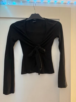 Svart omlottopp med knyt framtill - Trendig svart omlottopp med långa ärmar och knyt framtill. Djup v-ringning och figurnära passform som ger en snygg siluett. 
