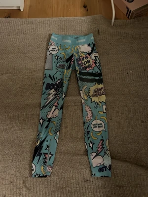 Stronger träningstights - Säljer ett par färgglada leggings från Stronger med coolt serietidningsinspirerat mönster i blått, gult, rosa och svart. Hög midja och stretchigt material som sitter skönt. Perfekta för dig som vill sticka ut med din outfit!