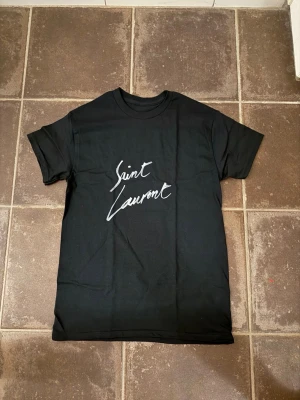 Saint Laurent t shirt - Riktigt fet och eftertraktad t shirt med saint Laurent loggan i storlek S! Helt nytt skick! Lite osäker på äktheten eftersom lappen i nacken saknas så därför är priset just nu på 499kr nypris runt 3000kr! Tveka inte med att ställa frågor! 