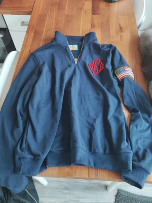 Epstein Hoodie (BRA KVALITE) - Mörkblå halvzip tröja från Carhartt med ribbade muddar och krage. På bröstet finns ett rött tryck med bokstäver och på ena ärmen sitter en patch med amerikanska flaggan. Perfekt för en avslappnad och sportig stil. Använd 1 gång och är väldigt skön.