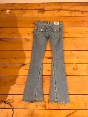 Blå bootcut jeans från True Religion i strl 23 - Säljer ett par ljusblå bootcut jeans från True Religion med klassiska kontrastsömmar och fickor med lock och knappar bak. Jeansen har låg midja och är tillverkade i denim med en lätt tvättad look. Perfekta för dig som gillar retrostil och detaljer. I strl 23 