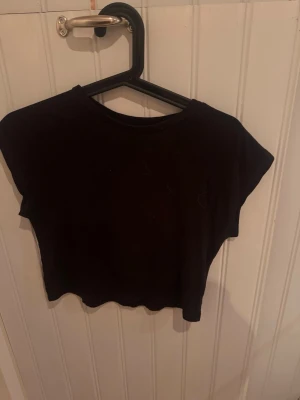 Svart croppad topp från H&M - En svart croppad topp från H&M med korta ärmar och rund hals. Enkel och stilren design som passar perfekt till jeans eller kjol. Tillverkad i mjuk bomull för en bekväm känsla.