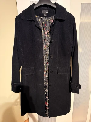 Svart manchesterrock från H&M - Snygg svart rock i manchestertyg från HM Rocken har knäppning framtill, två fickor och ett blommigt innerfoder som ger en cool kontrast. Klassisk krage och dekorativa knappar vid ärmsluten. Perfekt för dig som gillar stilrena och tidlösa plagg.