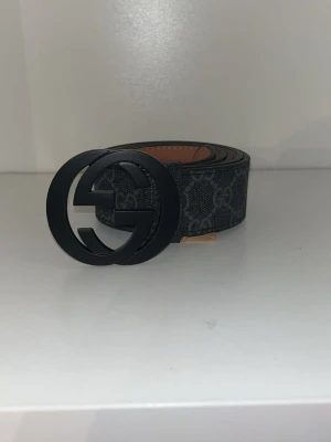 Svart GG-bälte från Gucci - Snyggt svart bälte från Gucci med klassiskt GG-mönster och stor svart GG-spänne. Bältet är gjort i skinn med detaljer i mörkgrått och har en exklusiv känsla. Perfekt för att lyfta vilken outfit som helst.