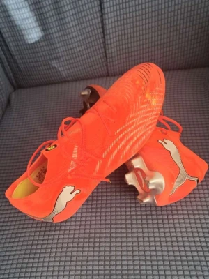Neonorange Puma fotbollsskor - Säljer ett par neonorange fotbollsskor från Puma med vita loggor på sidorna och silvriga dobbar. Skorna har snörning och en strumpliknande passform för extra komfort. Tillverkade i syntetmaterial som ger bra grepp och kontroll på planen.