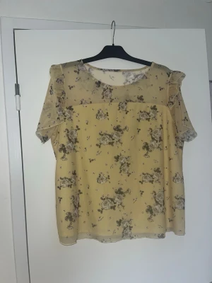 Gul blommig blus med volangärmar - Söt gul blus med små blommor i beige och grå toner. Blusen har transparenta volangärmar och axelparti i mesh, rund halsringning och rak passform. Perfekt för dig som gillar romantisk och feminin stil.