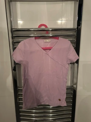 Ljuslila omlott t-shirt med v-ringning - Säljer en ljuslila t-shirt från Kappahl med omlottdetalj och v-ringad hals. T-shirten har korta ärmar och en liten rund läderpatch nertill. Perfekt för dig som gillar en enkel men ändå unik stil.