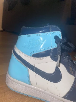 Air Jordan 1 Mid blå/vit - Säljer ett par Air Jordan 1 Mid sneakers i blått och vitt med svarta detaljer. Skorna har klassisk Jordan-logga på sidan, svart swoosh och svarta skosnören. Materialet är en mix av läder och syntet, och modellen har en hög siluett som ger en cool look.