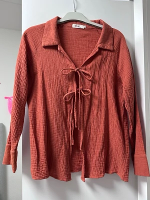 Rostfärgad blus med knyt  - Trendig rostfärgad blus i mjukt, krinklat bomullstyg. Blusen har långa ärmar, bred krage och två dekorativa knytband framtill. Perfekt för en avslappnad men stilsäker look.
