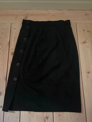 Svart kjol med knappar från H&M - Säljer nu en super elegant medellång kjol i ”sammet” material. Den sitter som en smäck och snatchar verkligen till i midjan. Den sitter gsnaks tajt runt kroppen. Super fin men kommer tyvärr ej till användning längre.💕