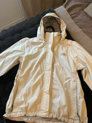 Vit vindjacka från The North Face - Snygg vit vindjacka från The North Face med huva och dragkedja framtill. Jackan har justerbar nederkant med dragsko och två sidofickor med dragkedja. Tillverkad i lätt och slitstarkt material, perfekt för blåsiga dagar.