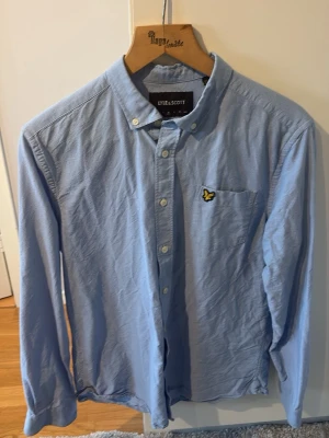 Blå skjorta från Lyle & Scott - Klassisk ljusblå skjorta från Lyle & Scott med button-down krage och bröstficka med den ikoniska gula loggan. Skjortan är långärmad, har normal passform och är tillverkad i mjuk bomull. Perfekt för en clean och stilren look.
