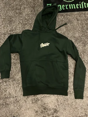 Mörkgrön hoodie från Pull&Bear - Mörkgrön hoodie från Pull&Bear med vit logga på bröstet. Klassisk känguruficka framtill och justerbar huva med dragsko. Tillverkad i mjukt material som känns skönt mot huden. Perfekt för dig som gillar streetwear och enkel stil.