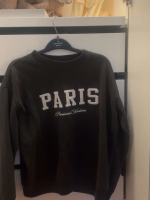 Svart sweatshirt med PARIS-tryck H&M - Cool svart sweatshirt från H&M med stort vitt PARIS-tryck och texten 'Parisienne Académie' på bröstet. Tröjan har rund halsringning och långa ärmar.