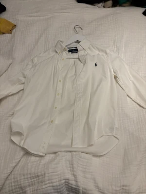 Vit skjorta från Ralph Lauren - Klassisk vit skjorta från Ralph Lauren med broderad logga på bröstet. Skjortan har lång ärm, knappar framtill och är tillverkad i bomull. Perfekt för en clean och stilren look. Hör av er vid mer info eller frågor! Storlek year 10-12